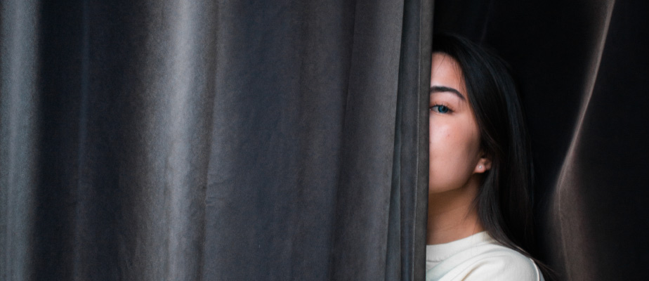 Brunette woman hides behind a gray curtain with only half her face showing due to embarrassment about her bad breath