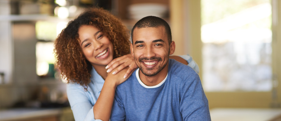 Dark-haired couple wearing blue shirts smile while sitting, the woman leaning on the man's shoulder