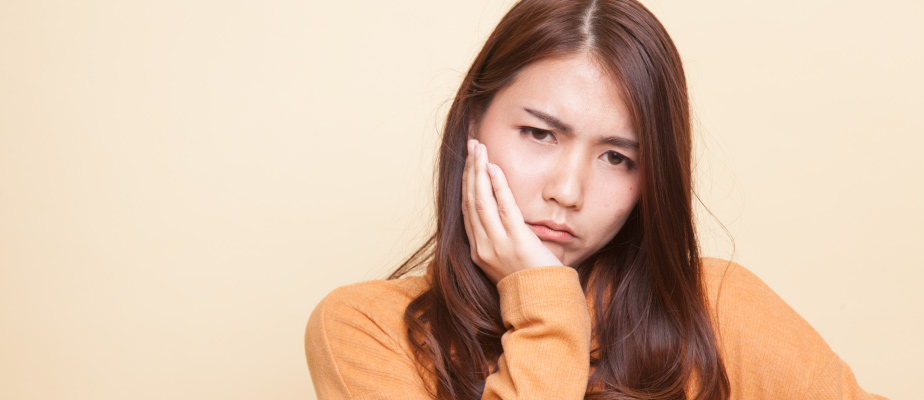 brunette woman wearing a yellow sweater cringes and touches her cheek in pain due to sensitive teeth