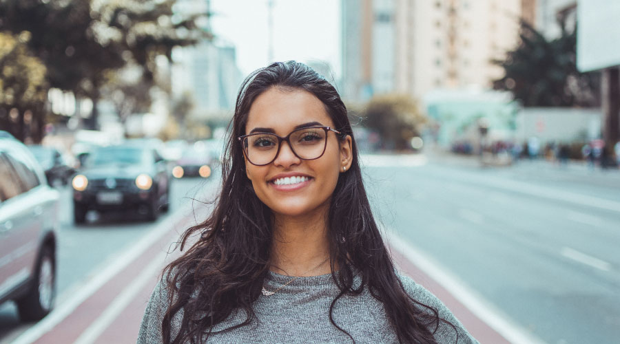 Brunette woman with strong enamel, glasses, and gray shirt smiles in a downtown of a busy city