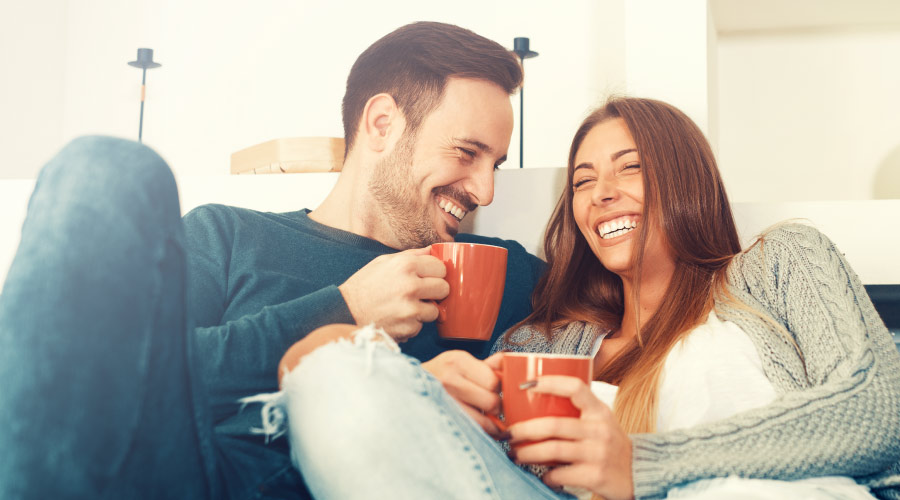 Man and woman with dental implants and veneers smile as they cuddle on the couch with orange mugs