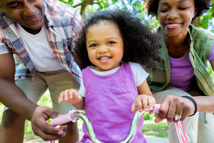 Little girl riding a trike with parents helping from behind.