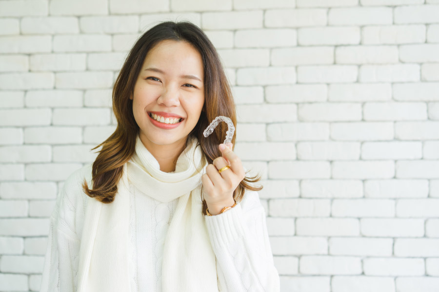 Young woman in front of a white brick wall is smiling and holding a ClearCorrect clear aligner.