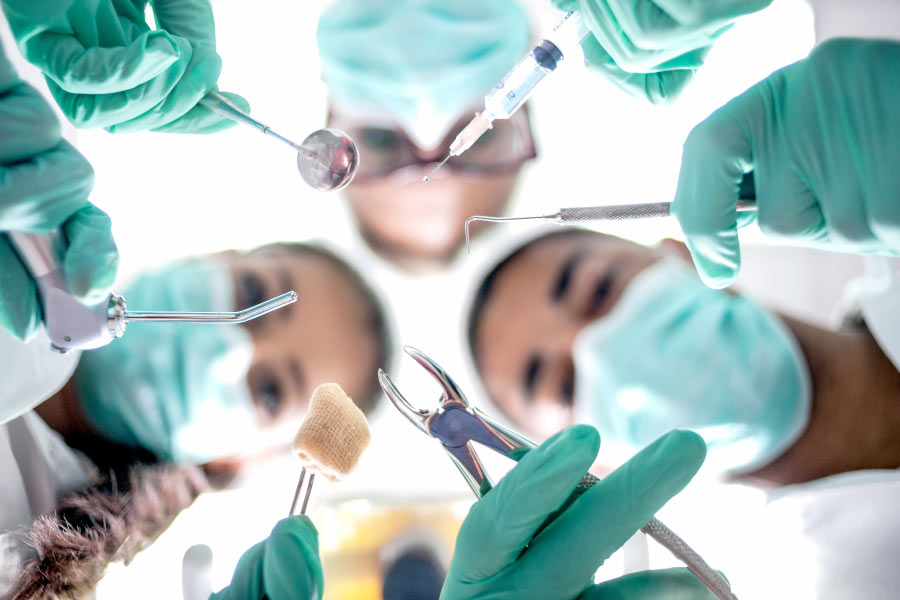 Masked dental professionals holding surgical tools are looking down on a patient ready for wisdom tooth extraction.