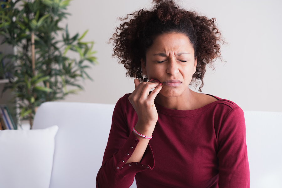 Black woman in a burgundy blouse cringes in pain and touches her cheek because she needs root canal therapy